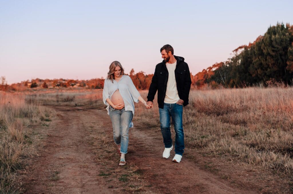Couple walking down a dirt track surrounded by dried grass and a blue fasing to pink sunset. Jeans and button up shirts